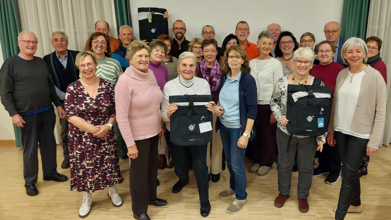Eine Gruppe von Erwachsenen, sowohl Männer als auch Frauen, sind für ein Foto versammelt. Eine Frau hält eine schwarze Tasche mit einem weißen Etikett. Der Raum hat einen Holzboden und eine weiße Wand.