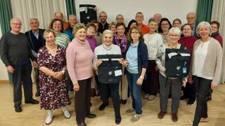 Eine Gruppe von Erwachsenen, sowohl Männer als auch Frauen, sind für ein Foto versammelt. Eine Frau hält eine schwarze Tasche mit einem weißen Etikett. Der Raum hat einen Holzboden und eine weiße Wand.
