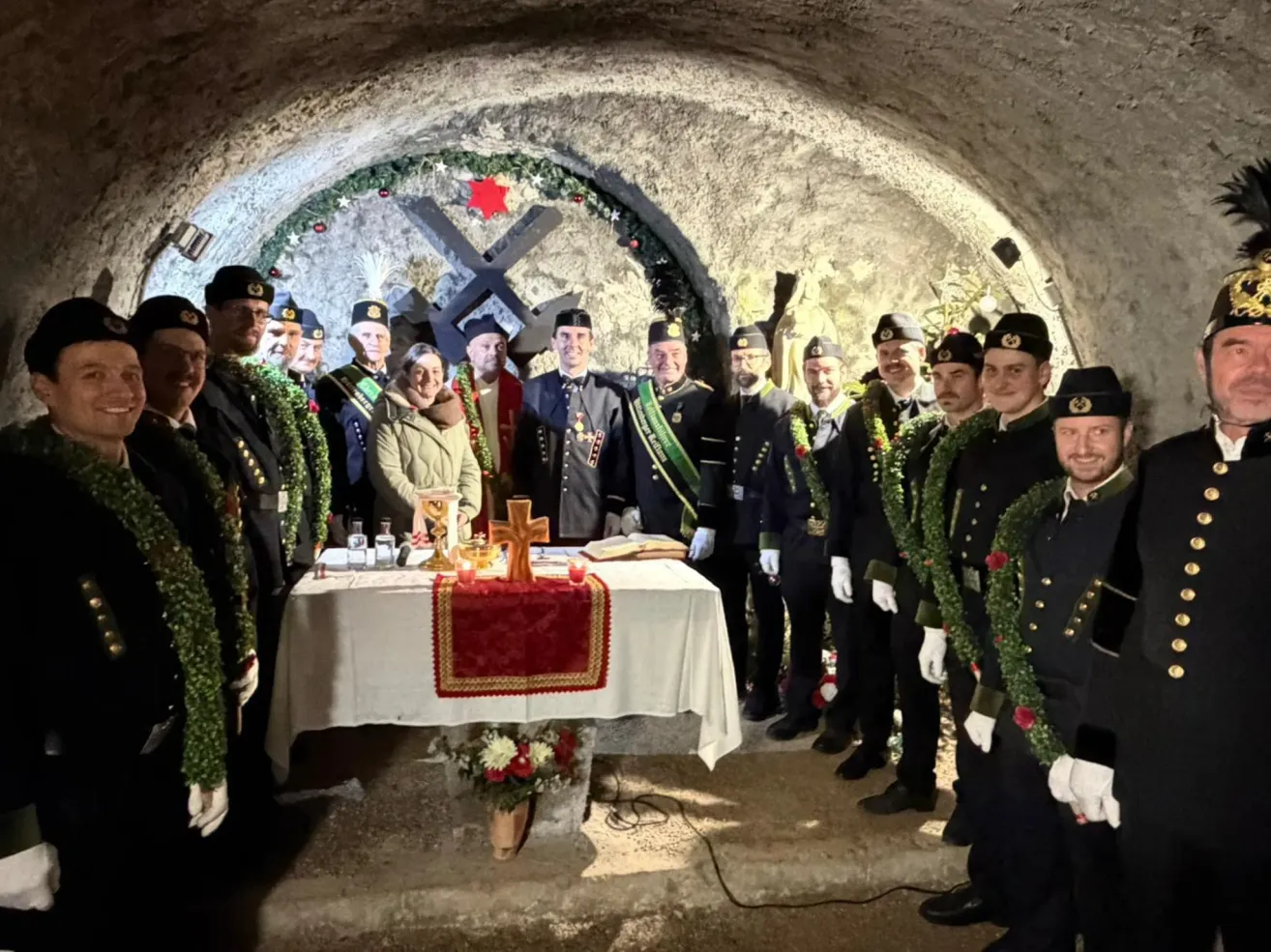 Eine Gruppe von Menschen in Uniform steht um einen Tisch mit einem Kreuz und Blumen herum, wahrscheinlich während einer Zeremonie. Sie sind mit Kränzen geschmückt.
