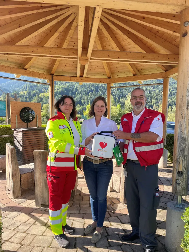 Drei Personen stehen in einem Pavillon, einer hält ein Defibrillator. Sie befinden sich in einer malerischen Gegend mit Bergen im Hintergrund.