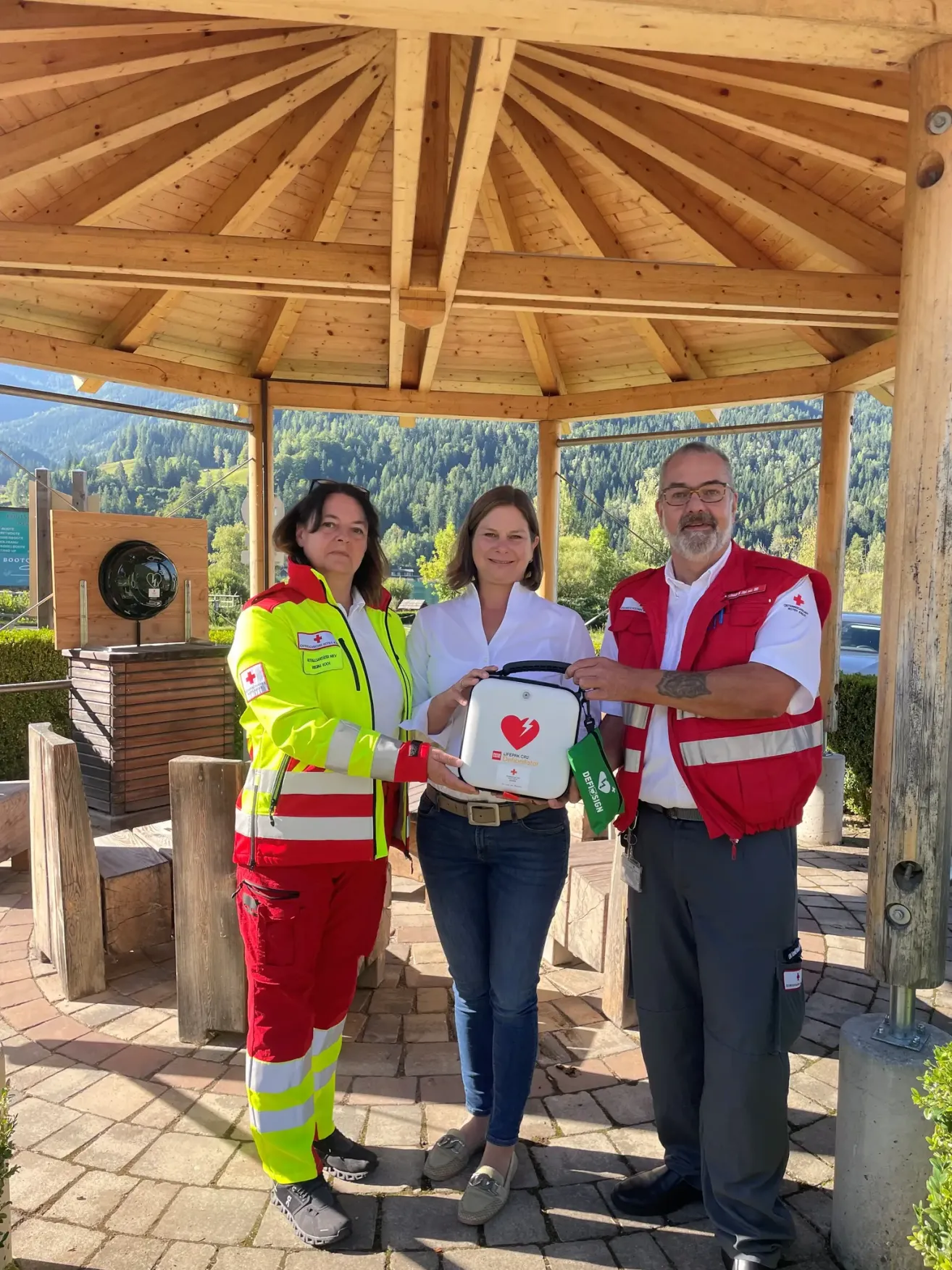 Drei Personen stehen in einem Pavillon, einer hält ein Defibrillator. Sie befinden sich in einer malerischen Gegend mit Bergen im Hintergrund.