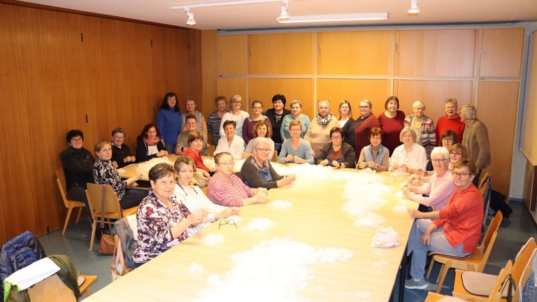 Eine Gruppe älterer Frauen sitzt um einen Tisch herum, einige tragen Brillen und Schals, und sie stricken. Der Tisch hat Wattebälle darauf.