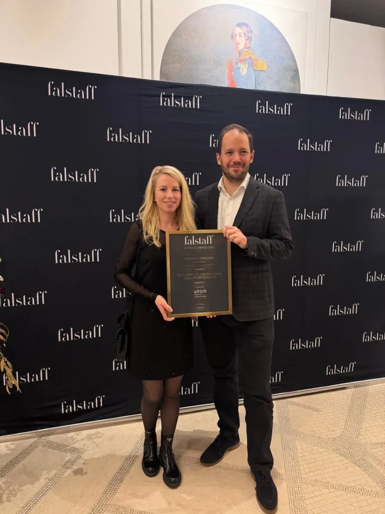 Ein Mann und eine Frau stehen vor einem schwarzen Hintergrund mit mehreren Falstaff-Logos. Sie halten einen gerahmten Preis und sehen glücklich aus.