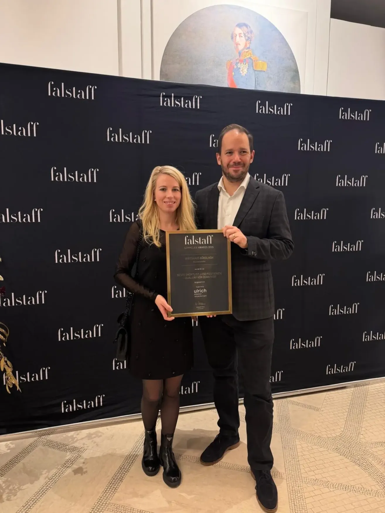 Ein Mann und eine Frau stehen vor einem schwarzen Hintergrund mit mehreren Falstaff-Logos. Sie halten einen gerahmten Preis und sehen glücklich aus.