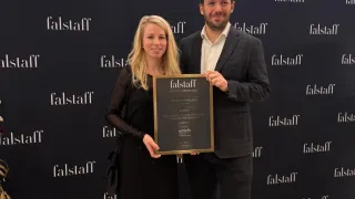 Ein Mann und eine Frau stehen vor einem schwarzen Hintergrund mit mehreren Falstaff-Logos. Sie halten einen gerahmten Preis und sehen glücklich aus.