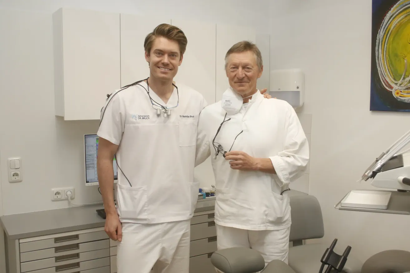 Zwei Zahnärzte in weißen Kitteln und Masken stehen in einer Zahnarztpraxis. Einer von ihnen lächelt, und der andere hat seine Hand auf der Schulter des anderen. Hinter ihnen steht ein Computermonitor auf einem Schreibtisch.