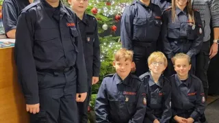 Eine Gruppe junger Menschen in Uniformen, wahrscheinlich Feuerwehrleute, posiert für ein Foto mit Erwachsenen um einen Weihnachtsbaum und ein großes Geschenk.