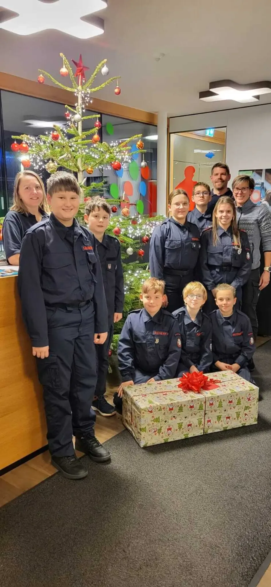 Eine Gruppe junger Menschen in Uniformen, wahrscheinlich Feuerwehrleute, posiert für ein Foto mit Erwachsenen um einen Weihnachtsbaum und ein großes Geschenk.