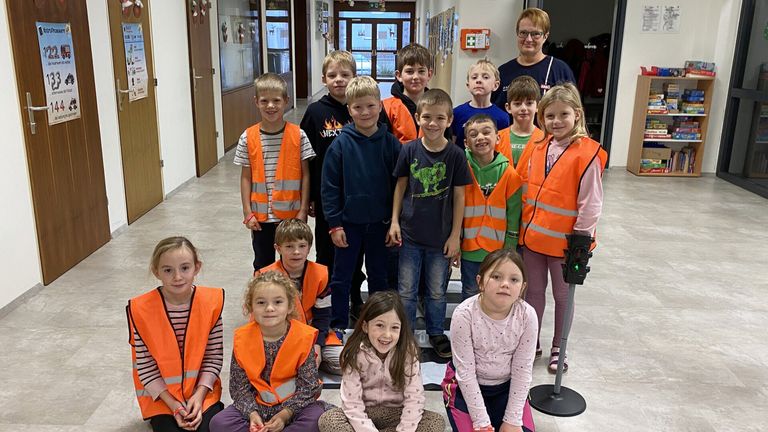Eine Gruppe von Kindern in Warnwesten posiert für ein Foto in einem Flur. Eine Frau steht hinter ihnen.