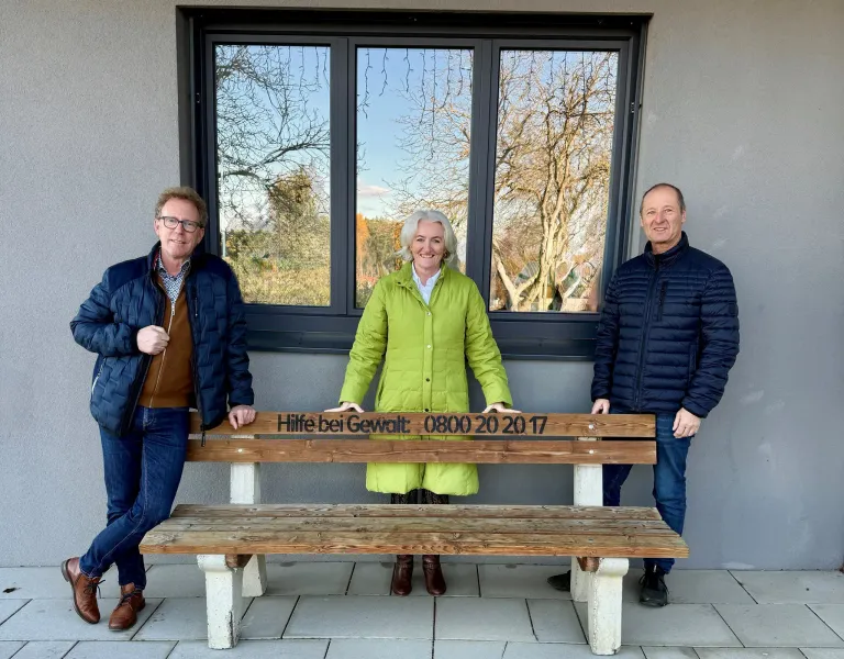 Drei Personen, zwei Männer und eine Frau, stehen neben einer Holzbank. Die Bank hat eine eingravierte Nachricht, und sie stehen vor einem Glasfenster.