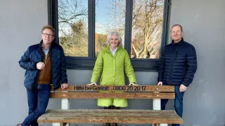 Drei Personen, zwei Männer und eine Frau, stehen neben einer Holzbank. Die Bank hat eine eingravierte Nachricht, und sie stehen vor einem Glasfenster.