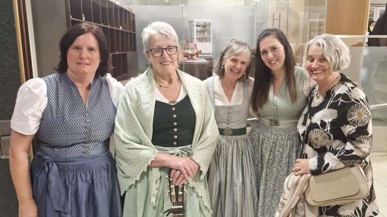 Vier Frauen lächeln in einem Raum, eine hält eine Gitarre. Sie tragen traditionelle Kleider, die Frau links trägt ein blaues Kleid. Hinter ihnen ist eine Glaswand, die einen kleinen Kühlschrank und Körbe zeigt.