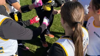 Eine Gruppe junger Frauen in Football-Uniformen ist auf einem Feld versammelt, eine hält einen Football, während ein Mann in einem Giants-Jacke zuschaut.