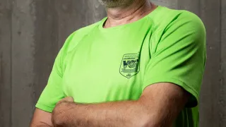 Ein Mann mit Bart und Schnurrbart in einem neongrünen Shirt steht mit verschränkten Armen vor einer Betonwand.