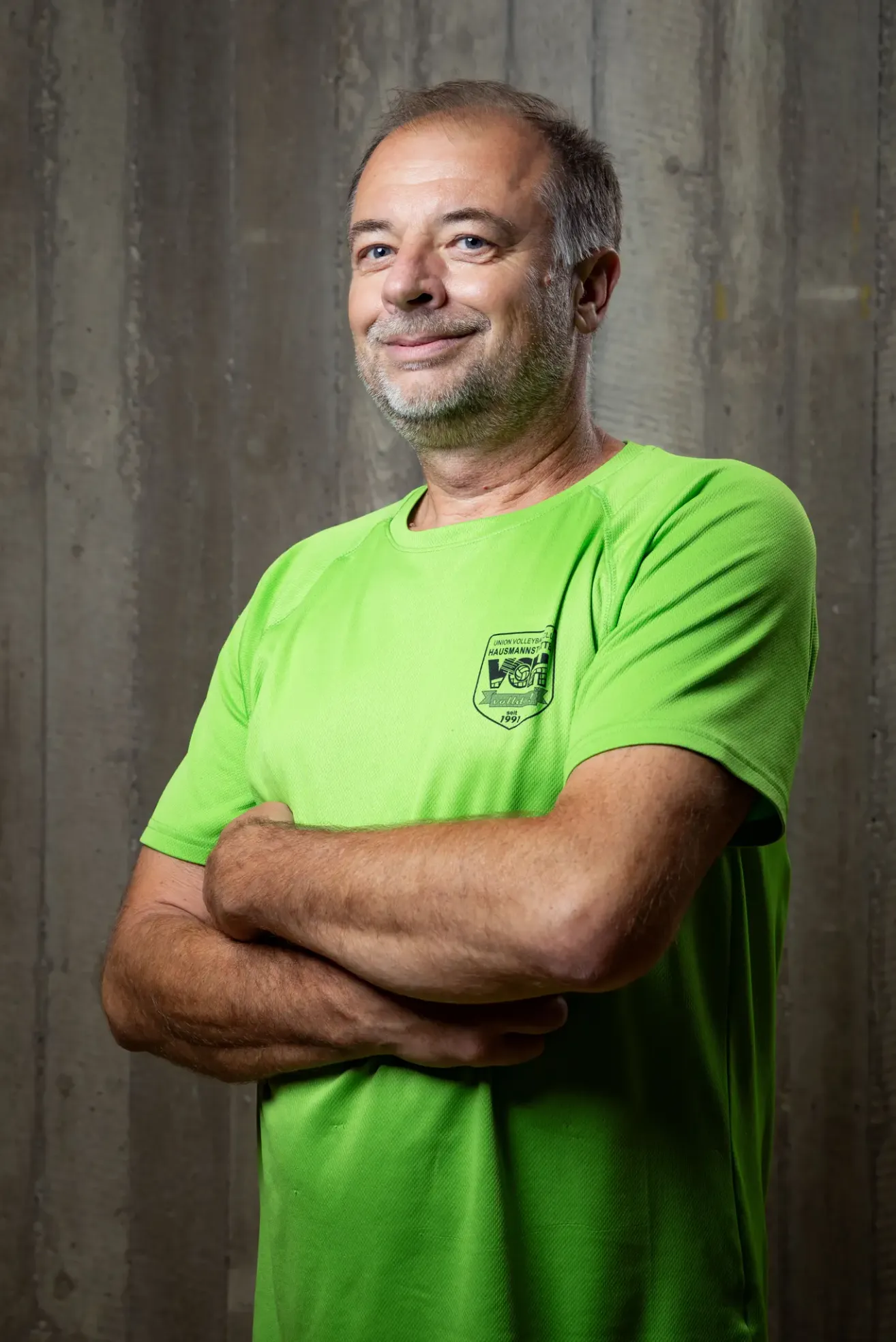 Ein Mann mit Bart und Schnurrbart in einem neongrünen Shirt steht mit verschränkten Armen vor einer Betonwand.