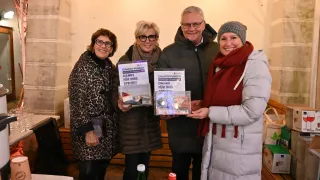 Vier Erwachsene stehen lächelnd und halten jeweils einen durchsichtigen Behälter mit Geld darin. Zwei Frauen tragen Schals und Brillen, und ein Mann trägt eine Mütze und eine Brille. Sie stehen vor einer beigen Wand mit einem Holzregal.