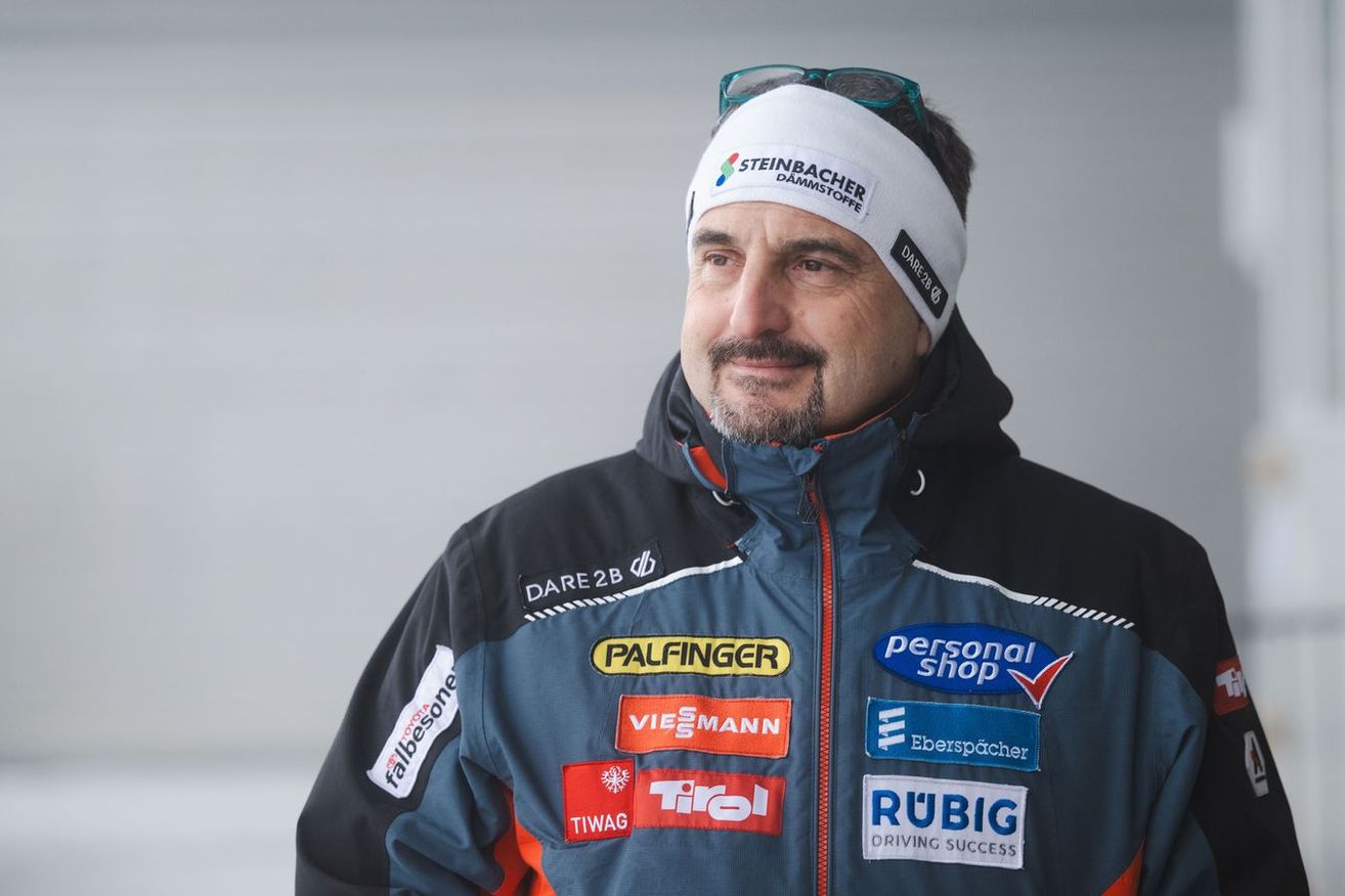 Ein Mann in einer Jacke mit mehreren Sponsorenlogos, darunter 'Personal Shop', 'Dare 2b', 'Pfeifener', 'Fallbeone', 'Tiwag', 'Rubig' und 'Eberspacher'. Er trägt auch eine Steinbacher Damstoffe-Stirnband.