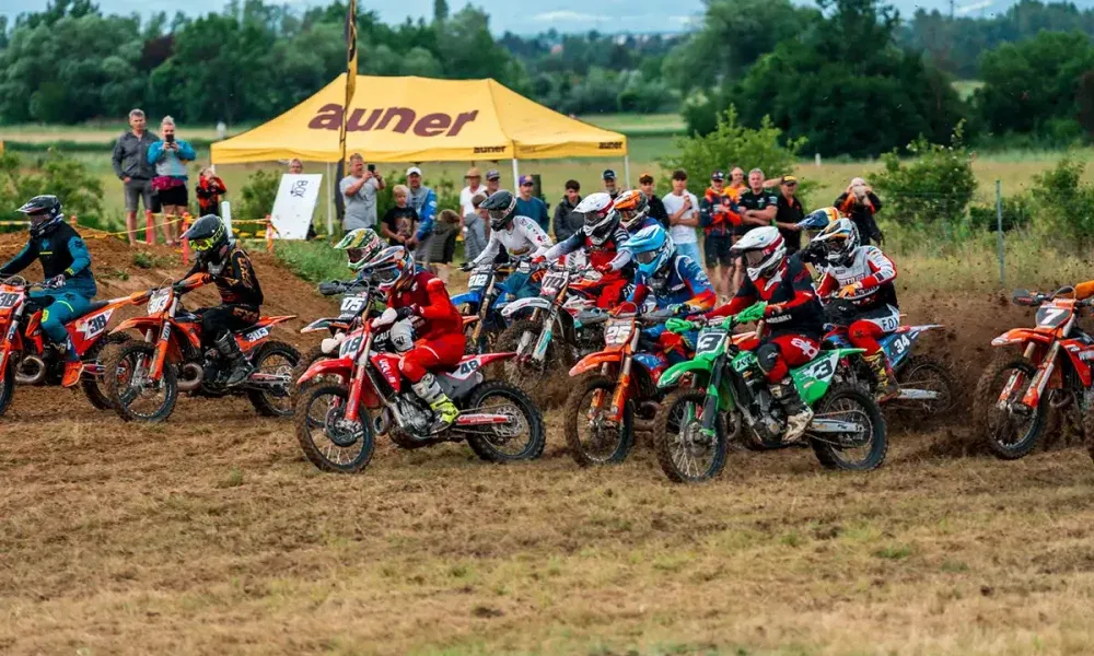 Motorradrennfahrer nehmen an einem Rennen auf einer Erdbahn teil, mit einem gelben Zelt und Zuschauern im Hintergrund. Die Fahrer tragen Helme und Rennanzüge, und die Motorräder sind nummeriert.
