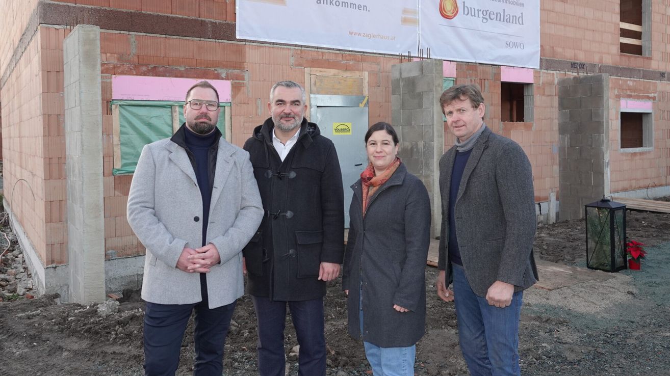 Vier Personen stehen vor einer Baustelle. Hinter ihnen befindet sich eine Ziegelmauer mit einem Banner. Ein Mann trägt eine Brille und einen grauen Mantel.