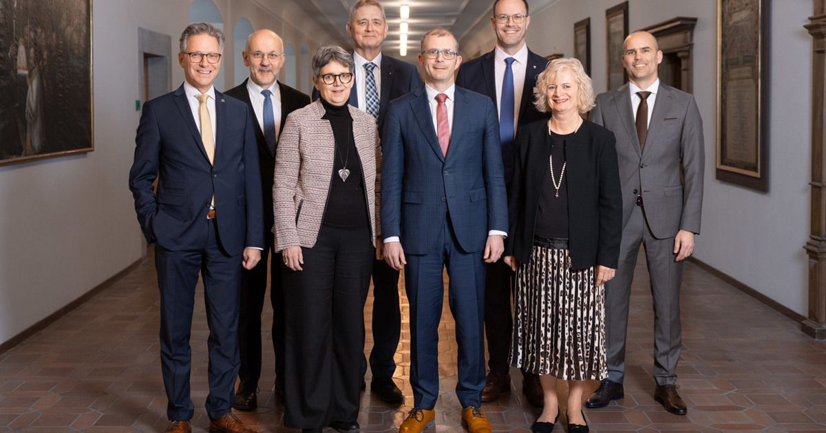 Eine Gruppe von Fachleuten in formeller Kleidung steht in einem Flur, lächelt und schaut in die Kamera.