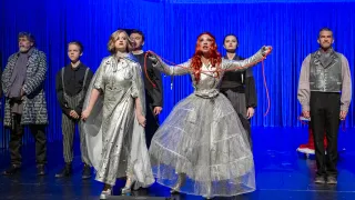 Eine Gruppe von Darstellern auf einer Bühne mit blauem Hintergrund. Eine Frau in silberner Kleidung hält eine rote Schnur, während andere zusehen.