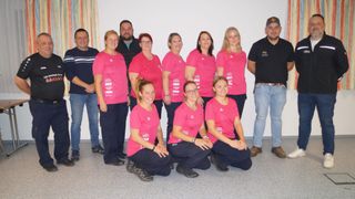 Eine Gruppe von Menschen in rosa T-Shirts posiert für ein Foto. Einige stehen, andere sitzen. Es gibt zwei Männer in schwarzen Hemden, die an den Seiten stehen.