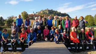 Eine Gruppe von Erwachsenen in verschiedenen Jackenfarben posiert für ein Foto auf einem Holzsteg vor einer Burg und Stadt.