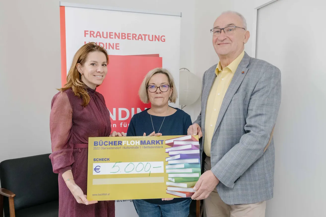 Drei Personen stehen vor einer weißen Wand mit einem roten Banner, das 'FRAUENBERATUNG INDINE' steht. Die Frau links hält einen Scheck über 5.000 € von Bucherer Flohmarkt.