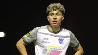 Ein junger Mann mit lockigem blondem Haar trägt ein Sporttrikot mit Logo und Nummern vor einem dunklen Hintergrund.