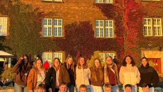 Eine Gruppe von Menschen in Jacken und Schals posiert vor einem Backsteingebäude, das mit Efeu und roten Blättern bedeckt ist. Der Hintergrund zeigt einen Baum und eine Steinmauer.
