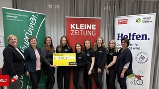 Eine Gruppe von Frauen, die schwarze Uniformen tragen, steht vor einem roten Banner. Sie halten einen gelben Scheck über 10.834,40 €, der einer Frau auf der linken Seite überreicht wird. Dahinter stehen drei Banner, darunter eines mit der Aufschrift 'kleine zeitung'.
