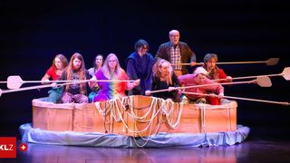 Eine Gruppe von Menschen ist auf der Bühne und rudert ein Pappboot, trägt verschiedene Kostüme und hält Paddel.