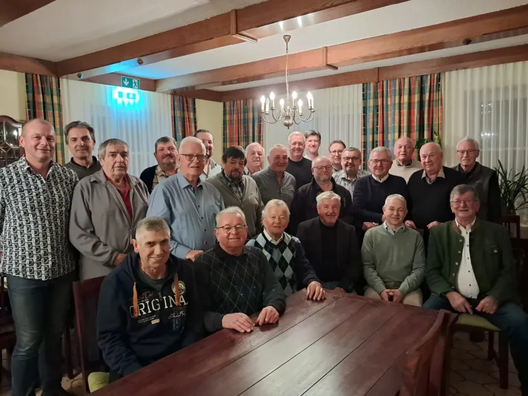 Eine Gruppe älterer Männer mit Brillen und Jacken posiert für ein Foto in einem Raum mit einem Holztisch, einer Kronleuchter und Vorhängen.