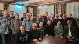 Eine Gruppe älterer Männer mit Brillen und Jacken posiert für ein Foto in einem Raum mit einem Holztisch, einer Kronleuchter und Vorhängen.