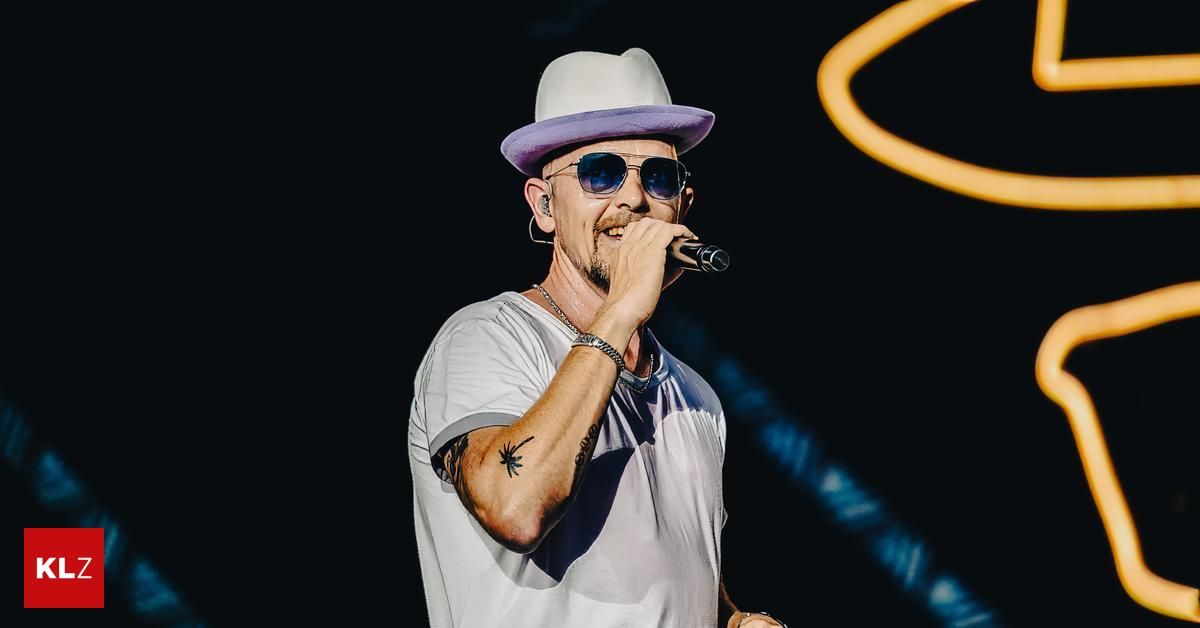 Ein Mann tritt auf der Bühne auf, trägt einen Hut und eine Sonnenbrille und singt in ein Mikrofon. Er hat eine Tätowierung am Arm.