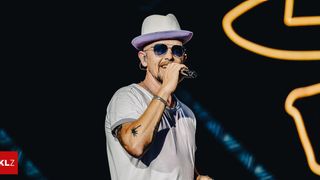 Ein Mann tritt auf der Bühne auf, trägt einen Hut und eine Sonnenbrille und singt in ein Mikrofon. Er hat eine Tätowierung am Arm.