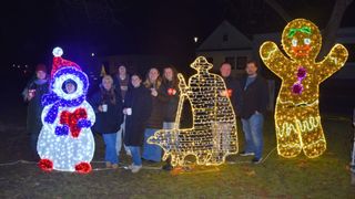 Eine Gruppe von Menschen in Wintermänteln steht um eine beleuchtete Weihnachtsdekoration eines Schneemanns und eines Mannes mit Hut. Sie halten Tassen und lächeln für die Kamera.