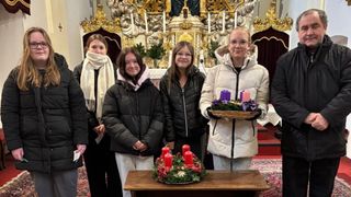 Vier Frauen stehen vor einem Altar mit Kerzen. Sie tragen Winterkleidung und eine hält einen Korb mit Kranz und Kerzen. Der Altar ist mit goldenen und weißen Elementen geschmückt.
