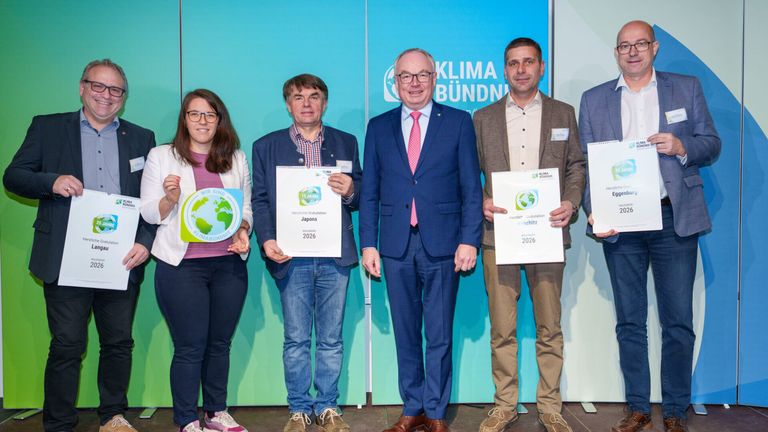 Fünf Personen stehen zusammen und halten Urkunden in der Hand, lächeln für ein Foto vor einem Hintergrund mit Klima Bündnis und grünen Erdbildern.