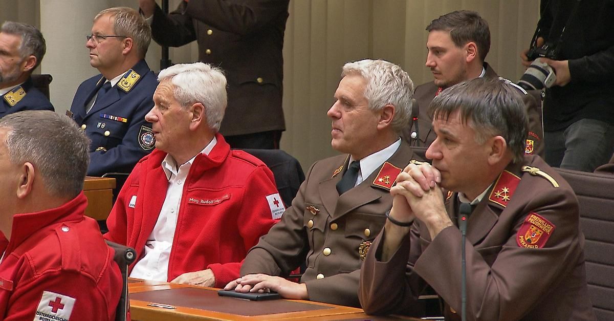 Fünf Männer in Militäruniformen und einem roten Jackett mit weißem Hemd sitzen an einem Tisch, möglicherweise in einer Besprechung. Ein Mann hält ein Tablet.