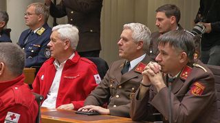 Fünf Männer in Militäruniformen und einem roten Jackett mit weißem Hemd sitzen an einem Tisch, möglicherweise in einer Besprechung. Ein Mann hält ein Tablet.