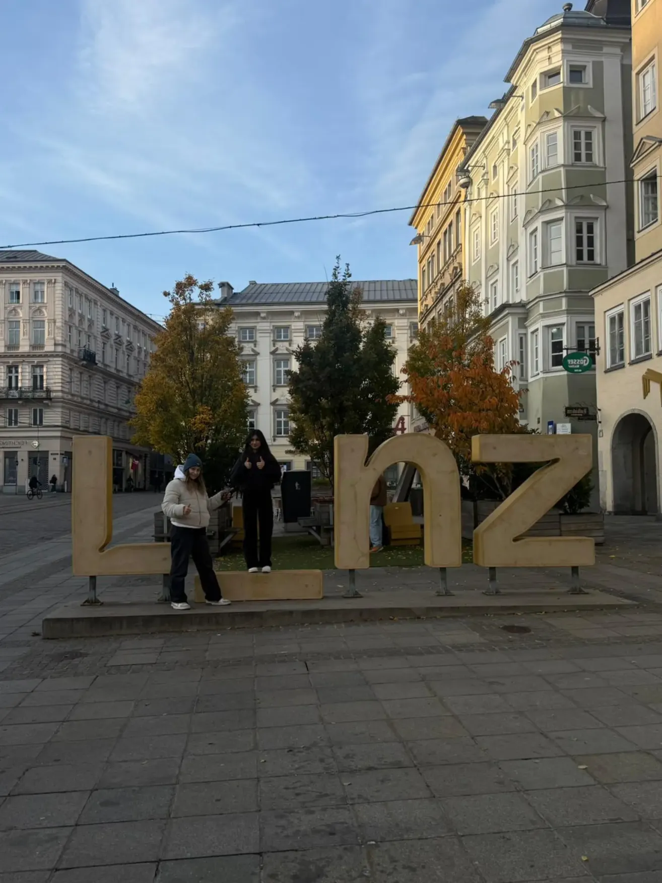 Zwei Frauen posieren für ein Foto vor einem großen Holzschild, das den Stadtnamen im Zentrum eines Stadtplatzes buchstabiert.