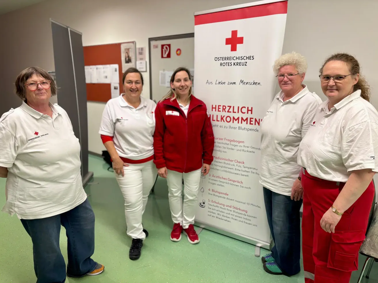 Fünf Frauen in weißen und roten Uniformen stehen vor einem Banner, das Herzlich Willkommen Aus Liebe zum Menschen besagt. Das Banner hat ein rotes Kreuz-Symbol und eine Liste von Anweisungen.