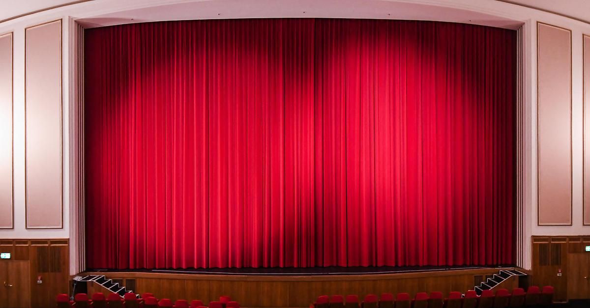 Eine leere Theaterbühne mit rotem Vorhang und roten Stühlen davor.