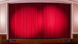 Eine leere Theaterbühne mit rotem Vorhang und roten Stühlen davor.