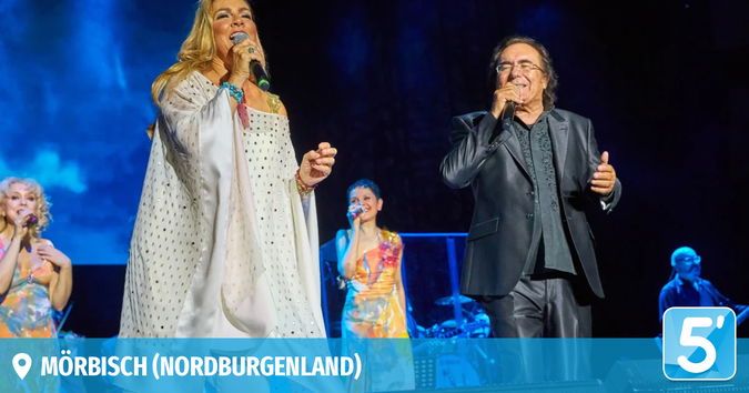 Zwei Künstler auf der Bühne, einer singt in ein Mikrofon und der andere gestikuliert. Dahinter sind weitere Sänger und eine Band zu sehen. Die Umgebung ist ein Konzert in Mörbisch, Nordburgenland, wie der Wasserzeichen verrät.