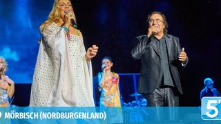 Zwei Künstler auf der Bühne, einer singt in ein Mikrofon und der andere gestikuliert. Dahinter sind weitere Sänger und eine Band zu sehen. Die Umgebung ist ein Konzert in Mörbisch, Nordburgenland, wie der Wasserzeichen verrät.