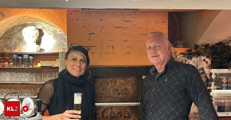 Ein Mann und eine Frau stehen lächelnd in einer Bar. Die Frau hält ein Glas Bier, und der Mann steht neben ihr. Hinter ihnen befindet sich eine Wand mit den Schildern Lania und Cosa.