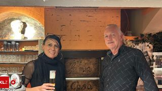 Ein Mann und eine Frau stehen lächelnd in einer Bar. Die Frau hält ein Glas Bier, und der Mann steht neben ihr. Hinter ihnen befindet sich eine Wand mit den Schildern Lania und Cosa.
