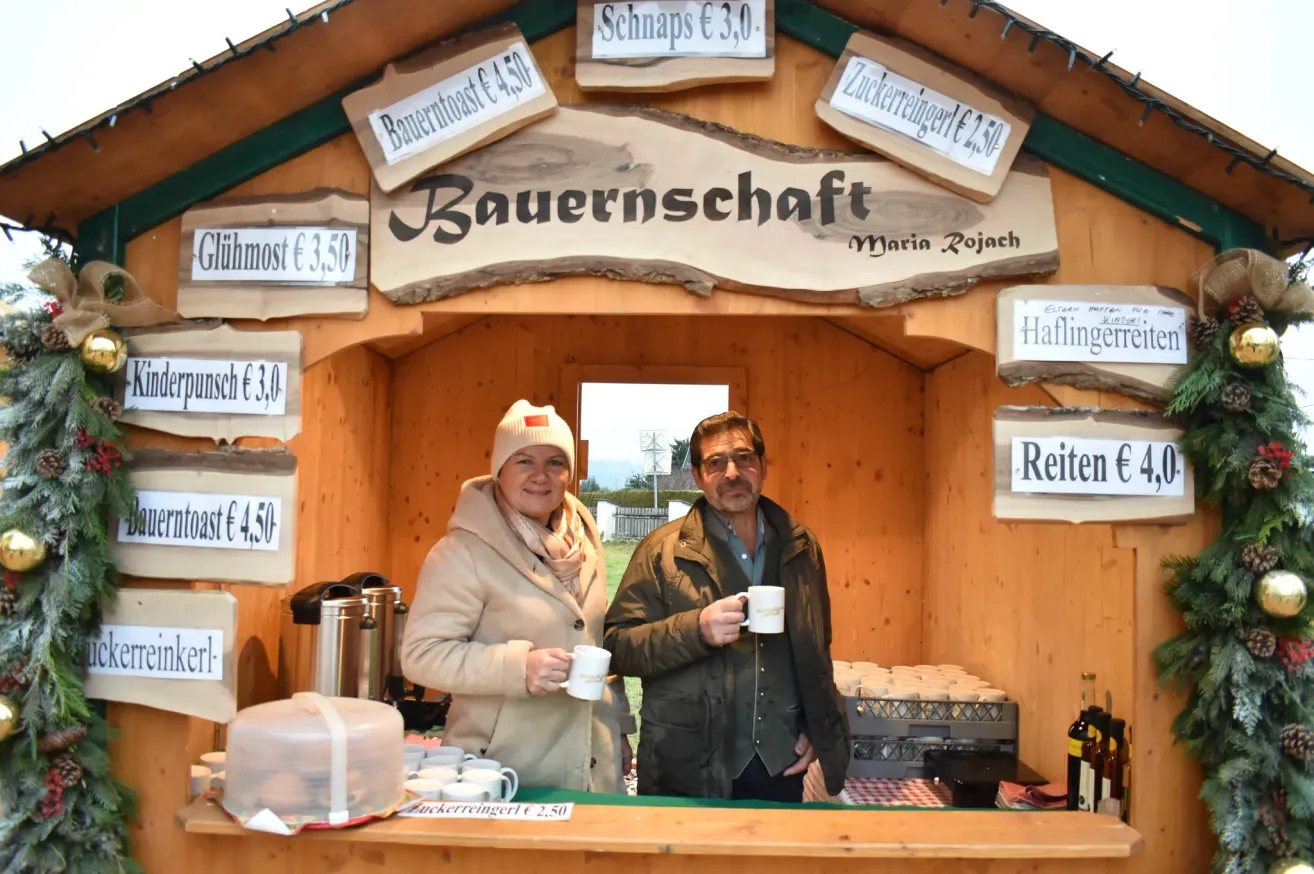 Zwei Personen stehen hinter einer Theke in einer Holzhütte mit ausgewiesenen Preisen an den Schildern. Eine Frau hält eine Tasse und ein Mann hält eine Tasse mit Logo.
